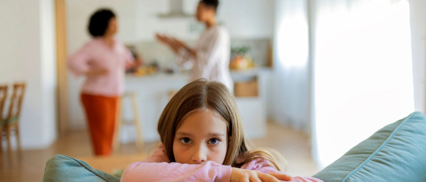 A young girl looking distressed on top of the couch with fighting adults blurred in the background.