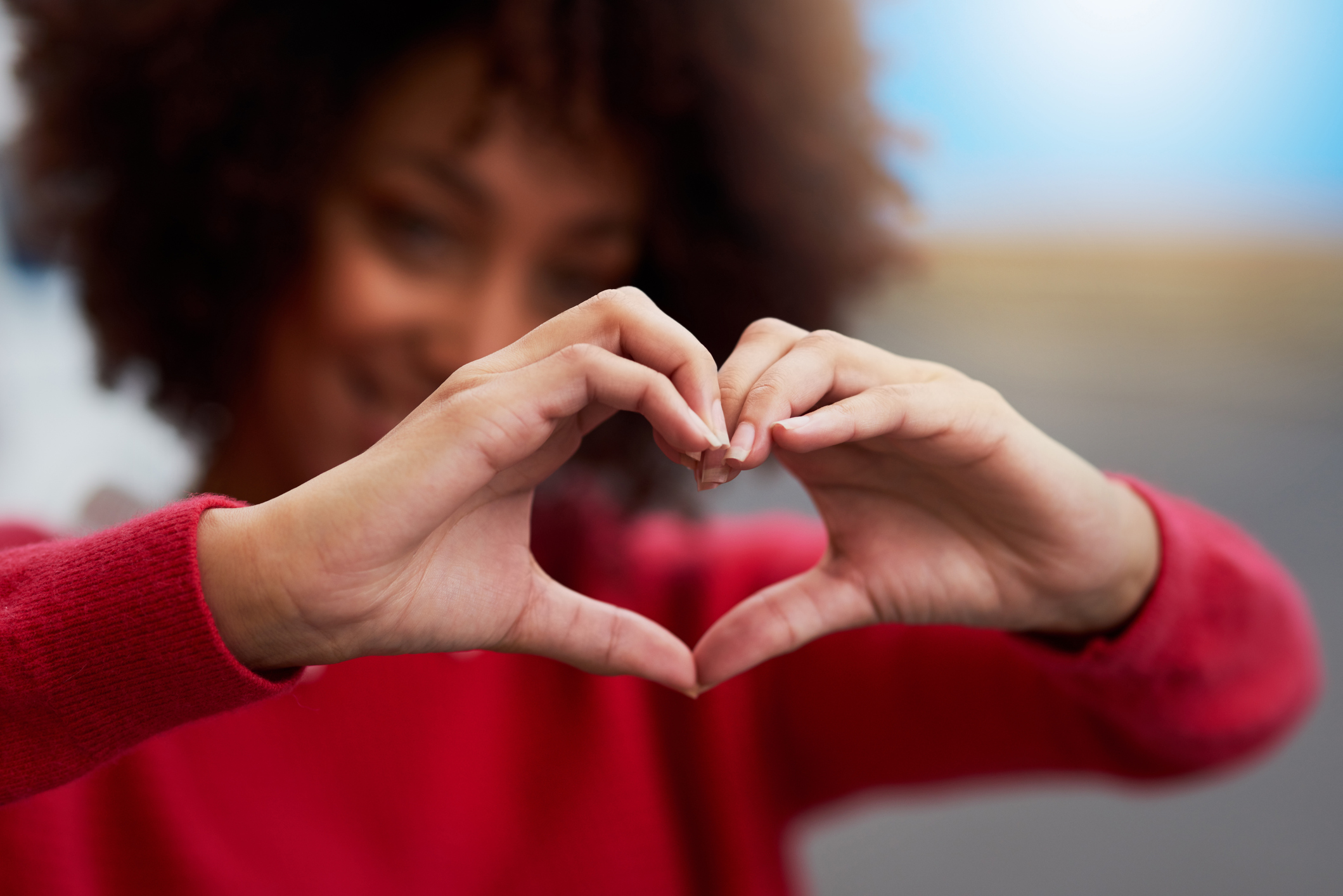 A person wearing a red sweater forms a heart shape with their hands, with their face softly blurred in the background.